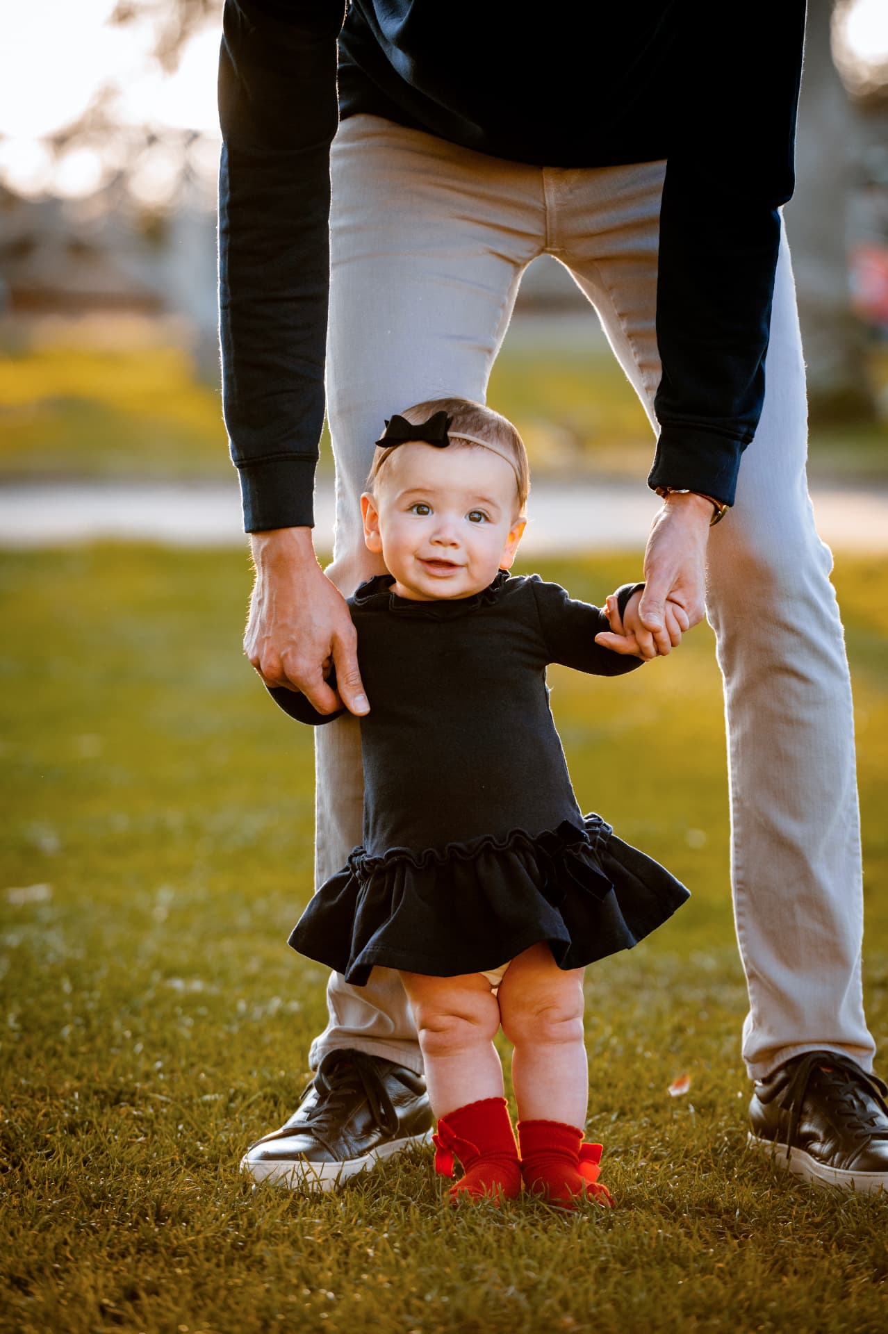Baby in a black ruffled dress and red booties holding her dad's hands