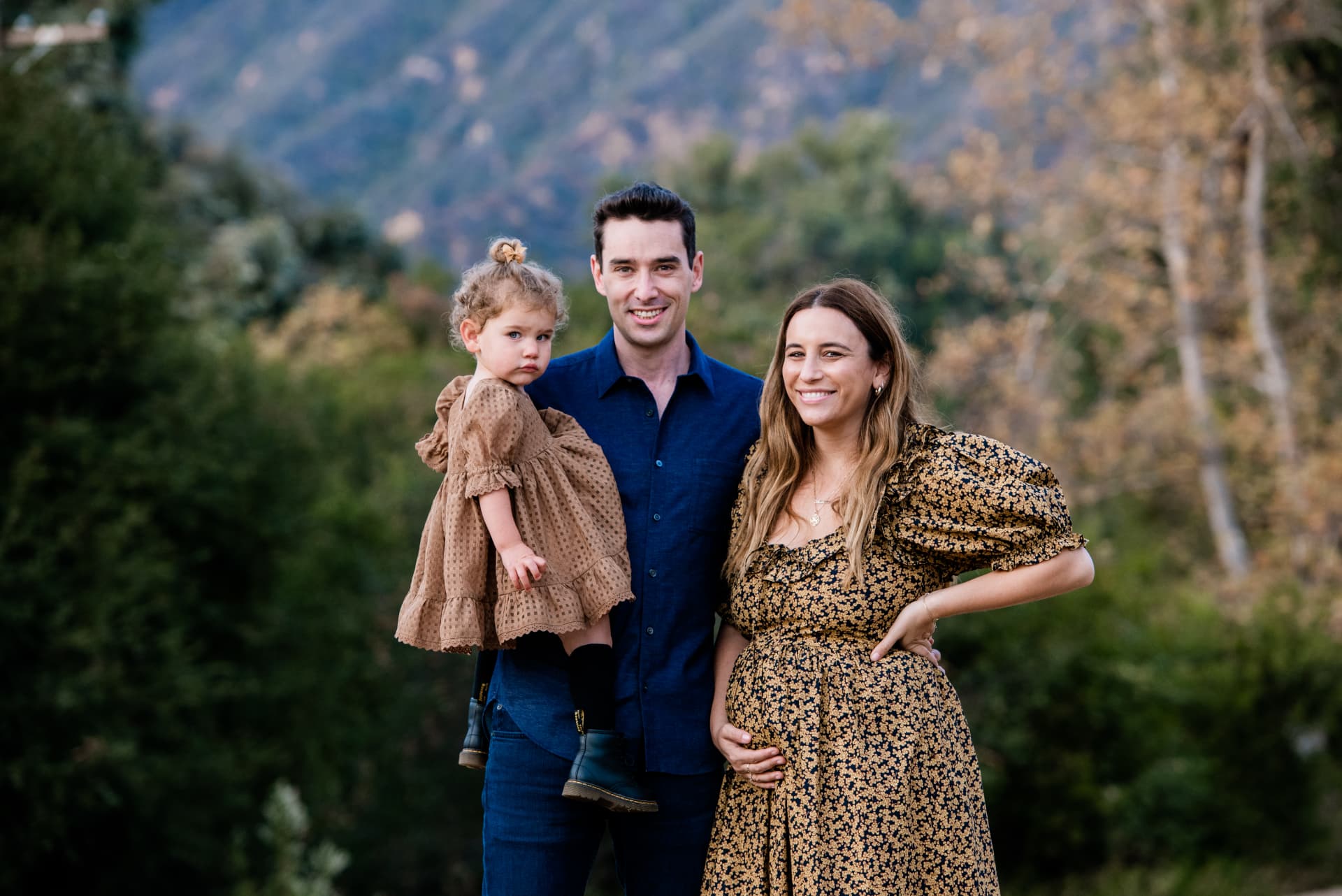 Family of three in denim and a floral dress in front of autumn hills