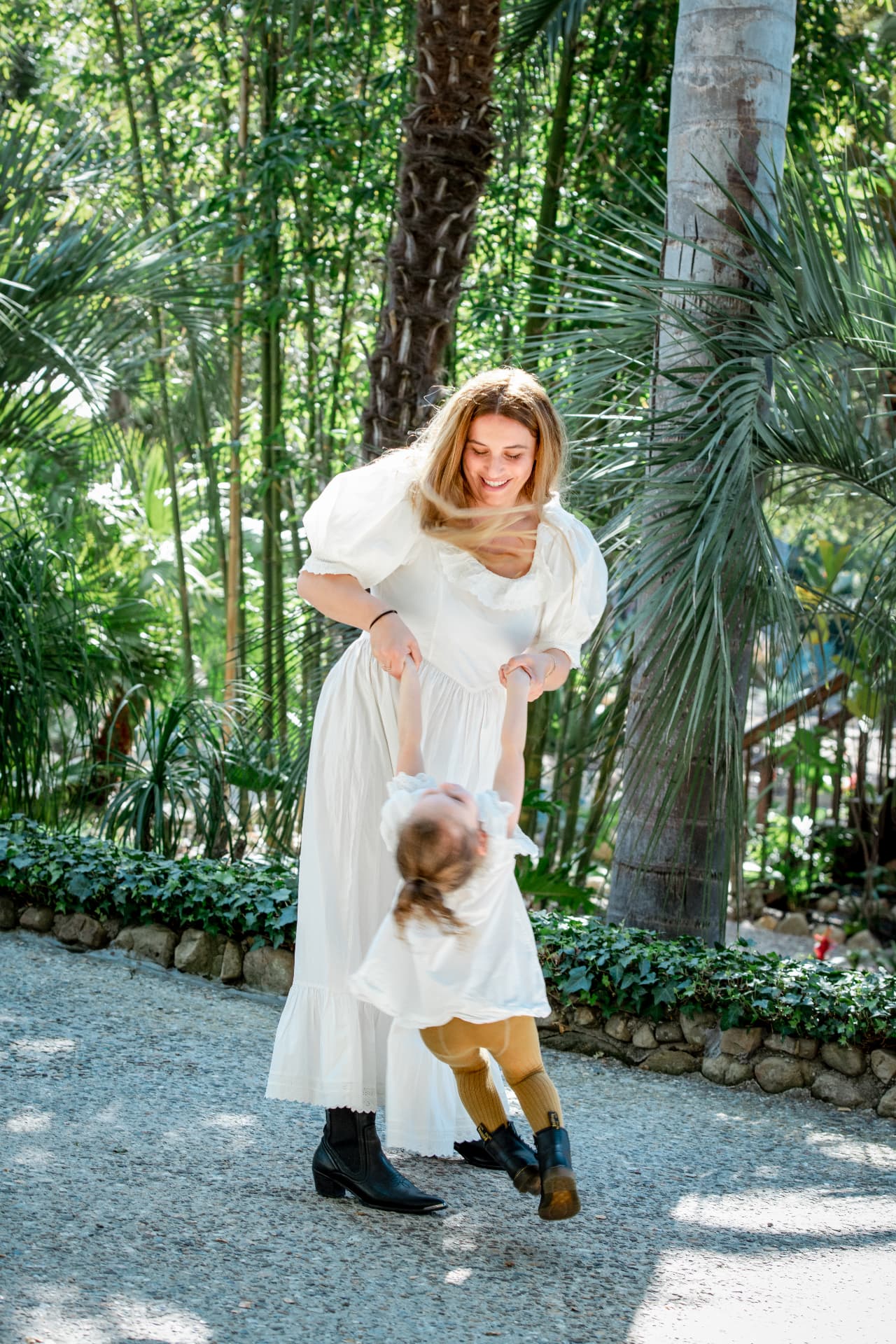 Mother in a white dress swinging her daughter on a garden path